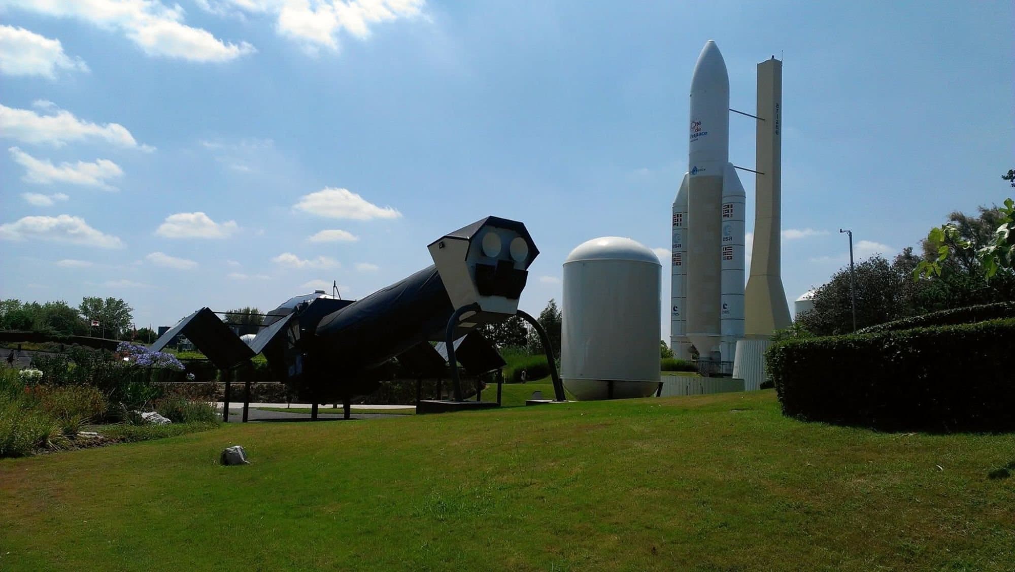 A Mir space station module exhibit in the foreground and an Ariane 5 replica under a cloudy sky