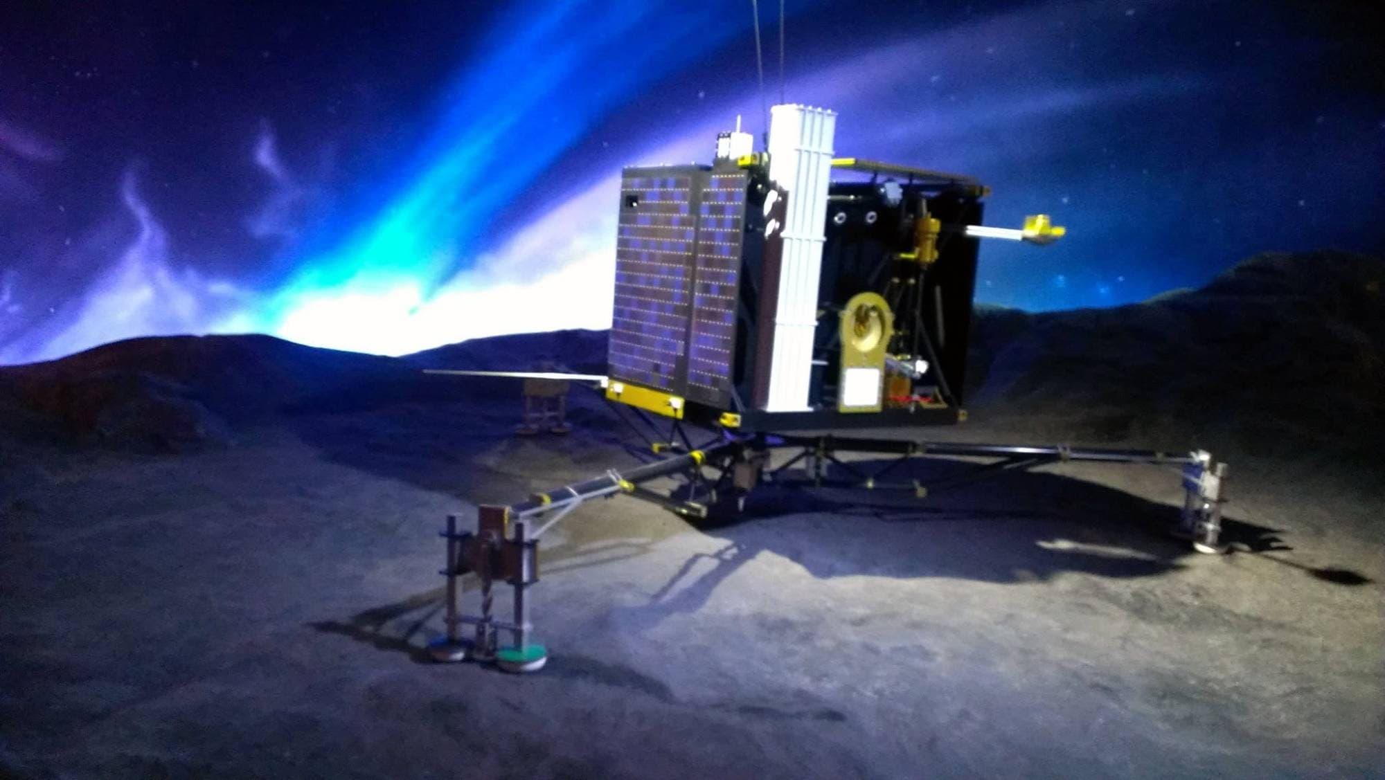A model of the Philae comet lander on a simulated comet surface under a blue glowing sky