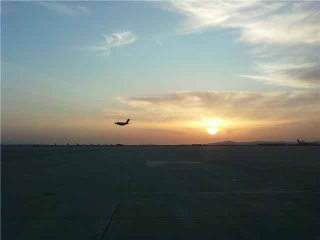A C-17 climbing away over a wide ramp at dusk, parked aircraft on the horizon